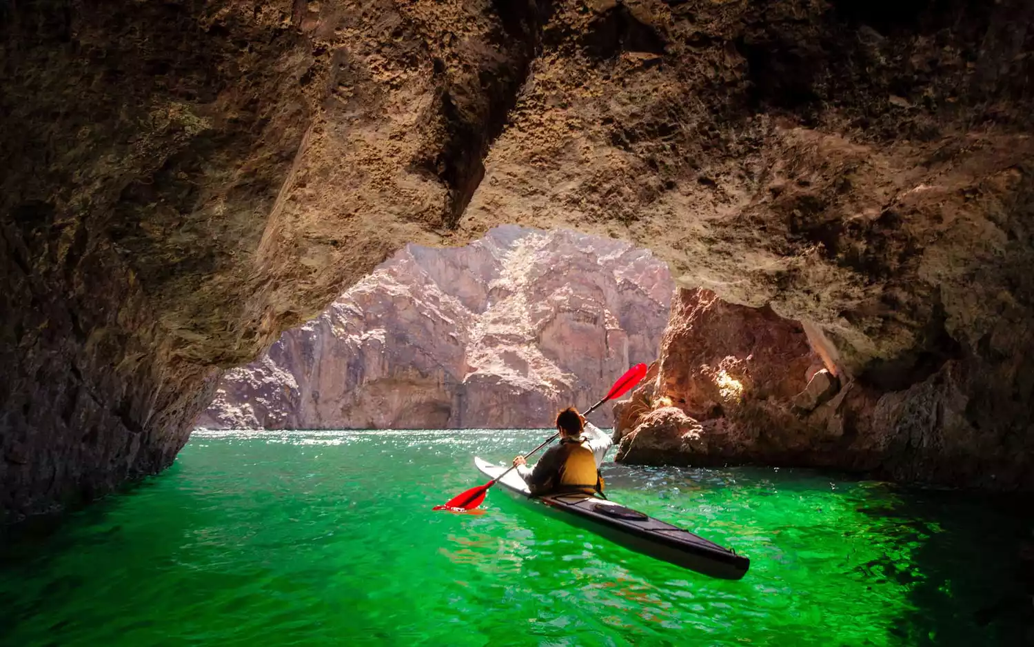 A kayaker navigating through the vibrant, glowing green water inside the mouth of a cave.