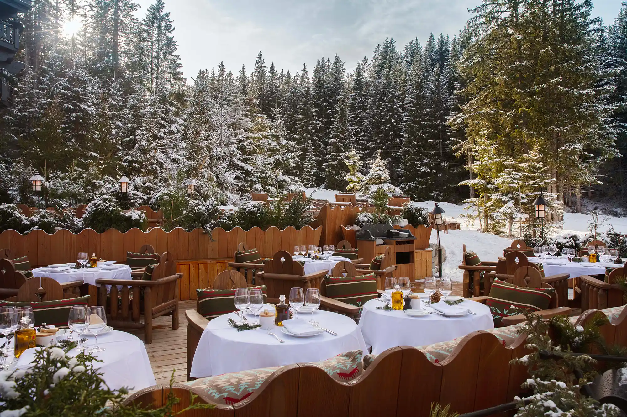 Outdoor dining deck with tables set for service against a backdrop of snowy pine trees.