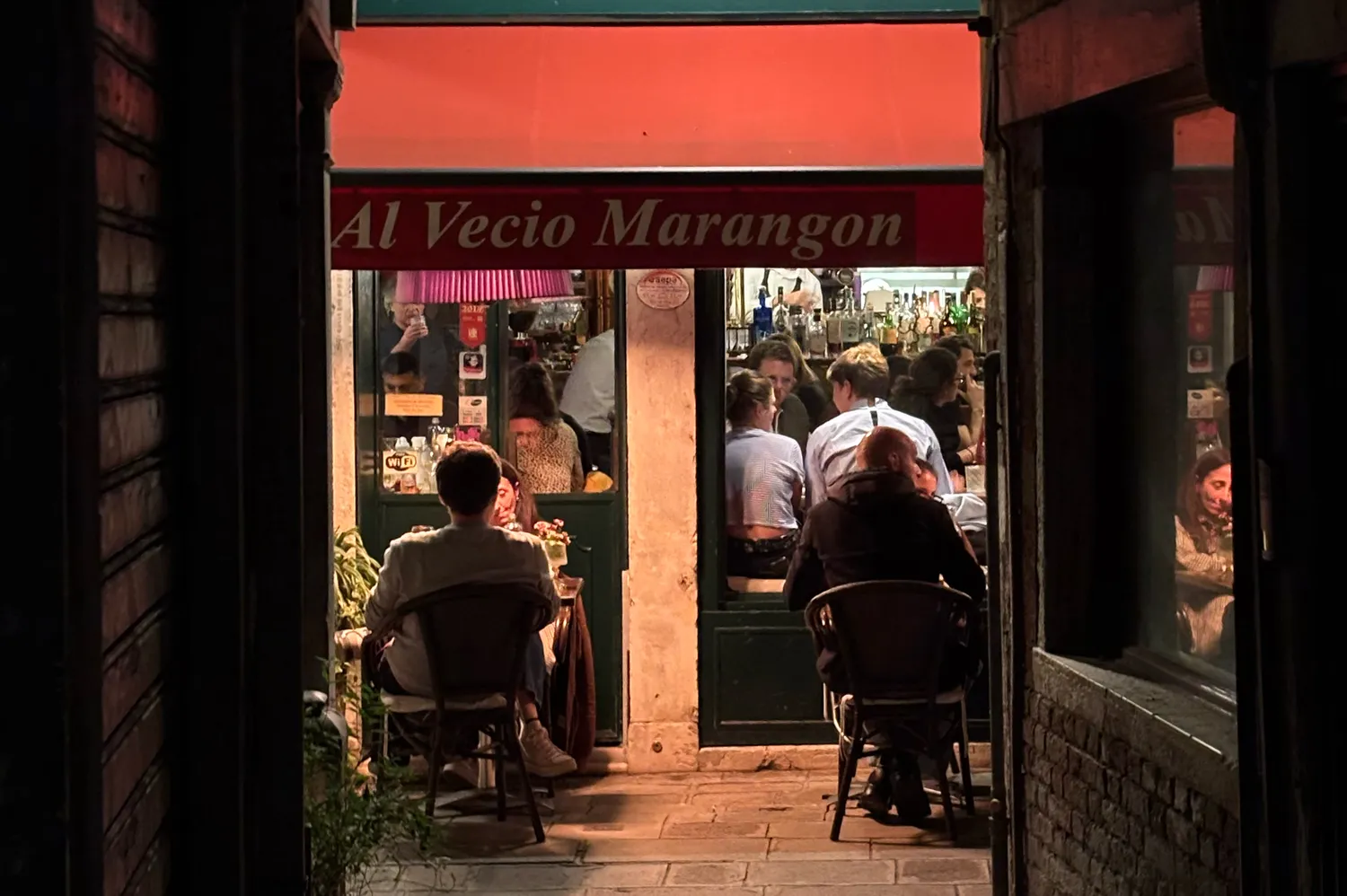 People dining and socializing in a lively evening atmosphere outside a traditional Venetian eatery.