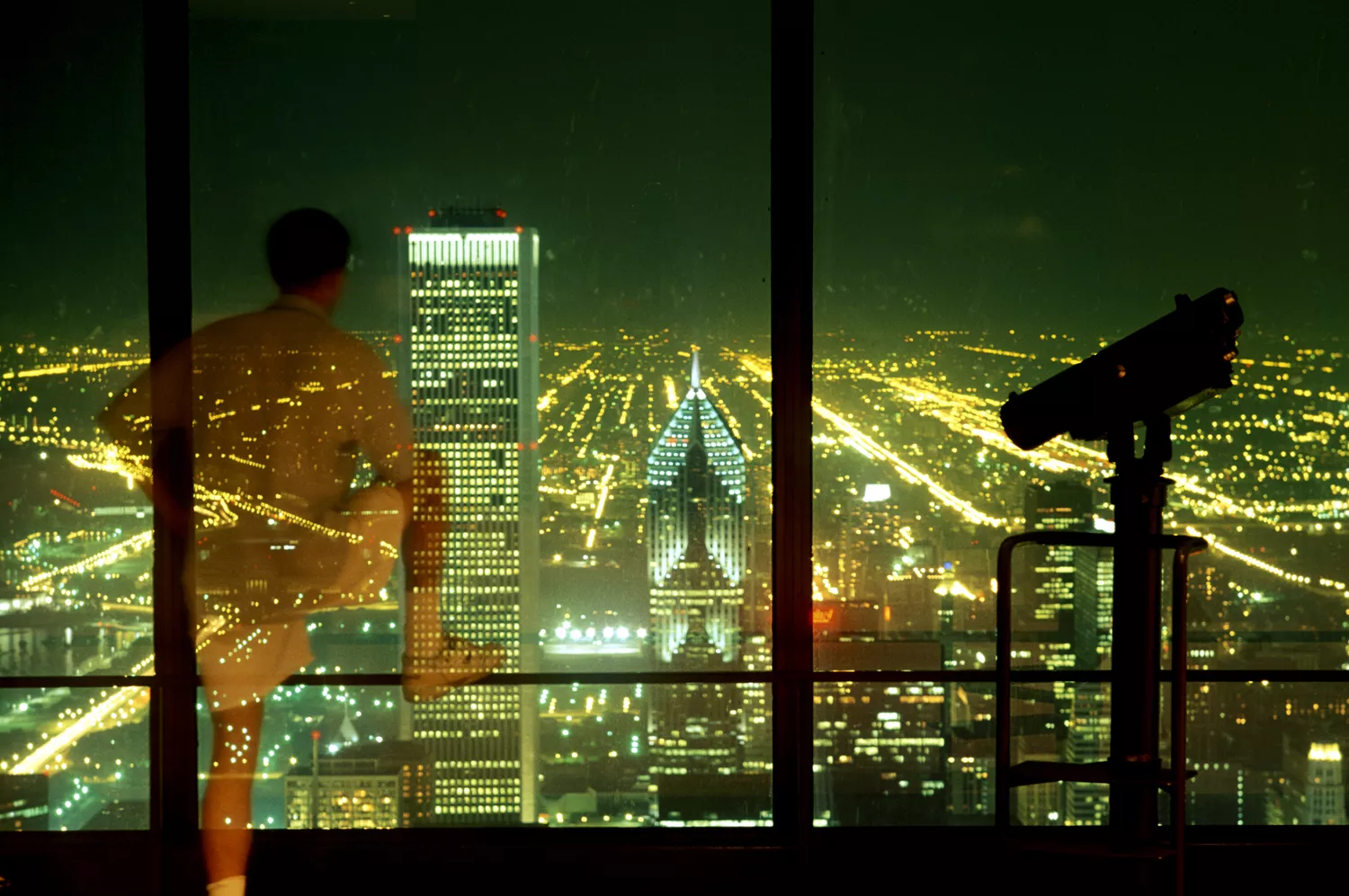 The Chicago city skyline at night seen through the windows of a high-altitude observation deck.