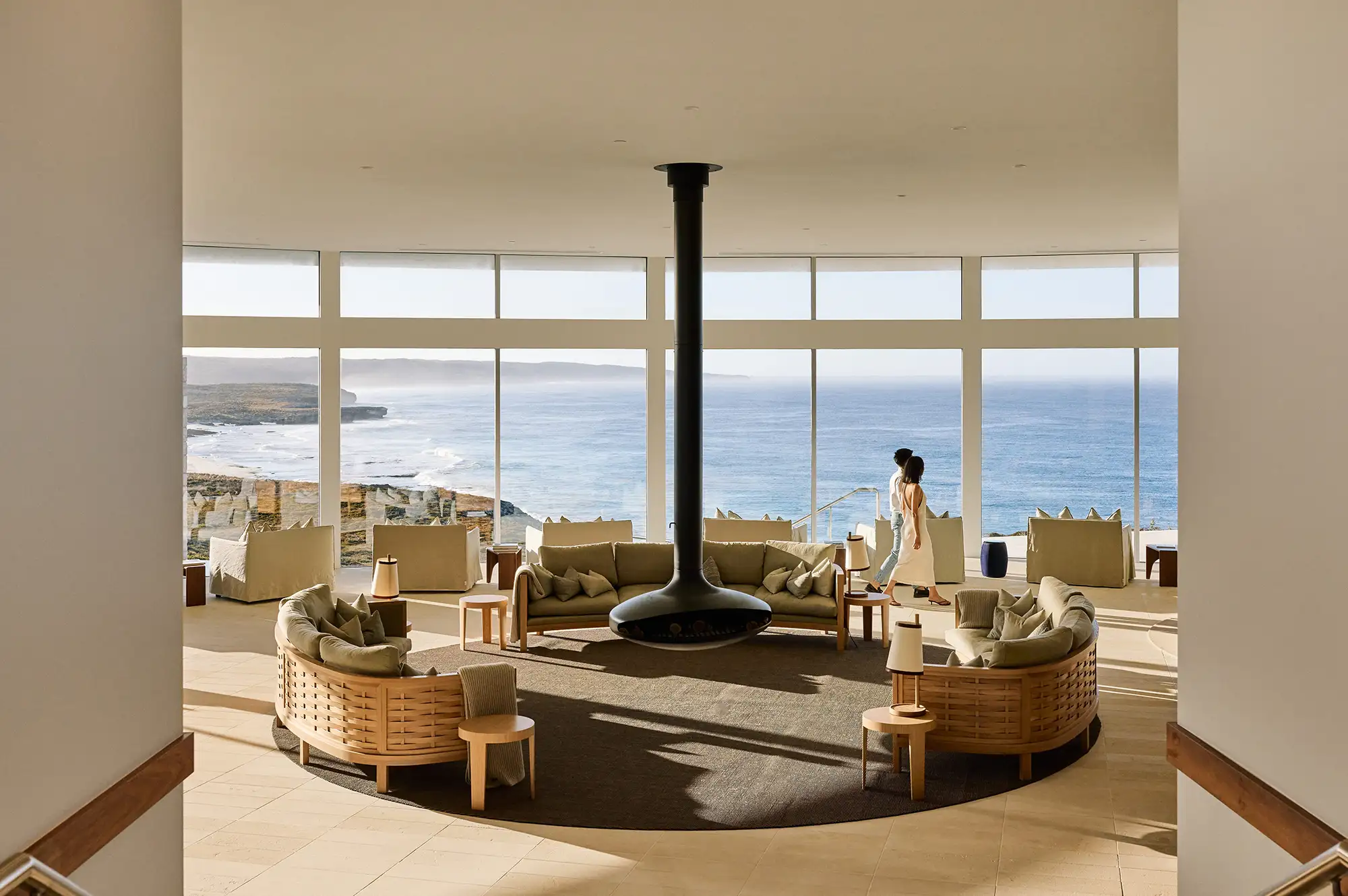 A modern hotel lounge with floor-to-ceiling windows overlooking the ocean and a central fireplace.