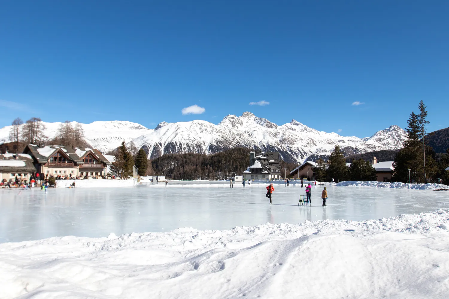 People ice skating on a frozen lake with snow-covered buildings and mountains in St. Moritz