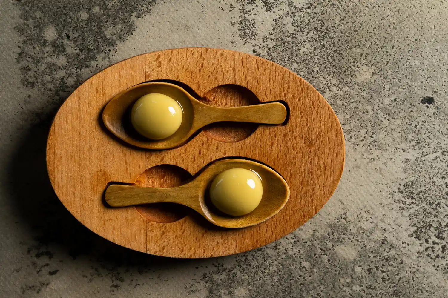Molecular gastronomy spherical servings presented on two wooden spoons on a textured base.