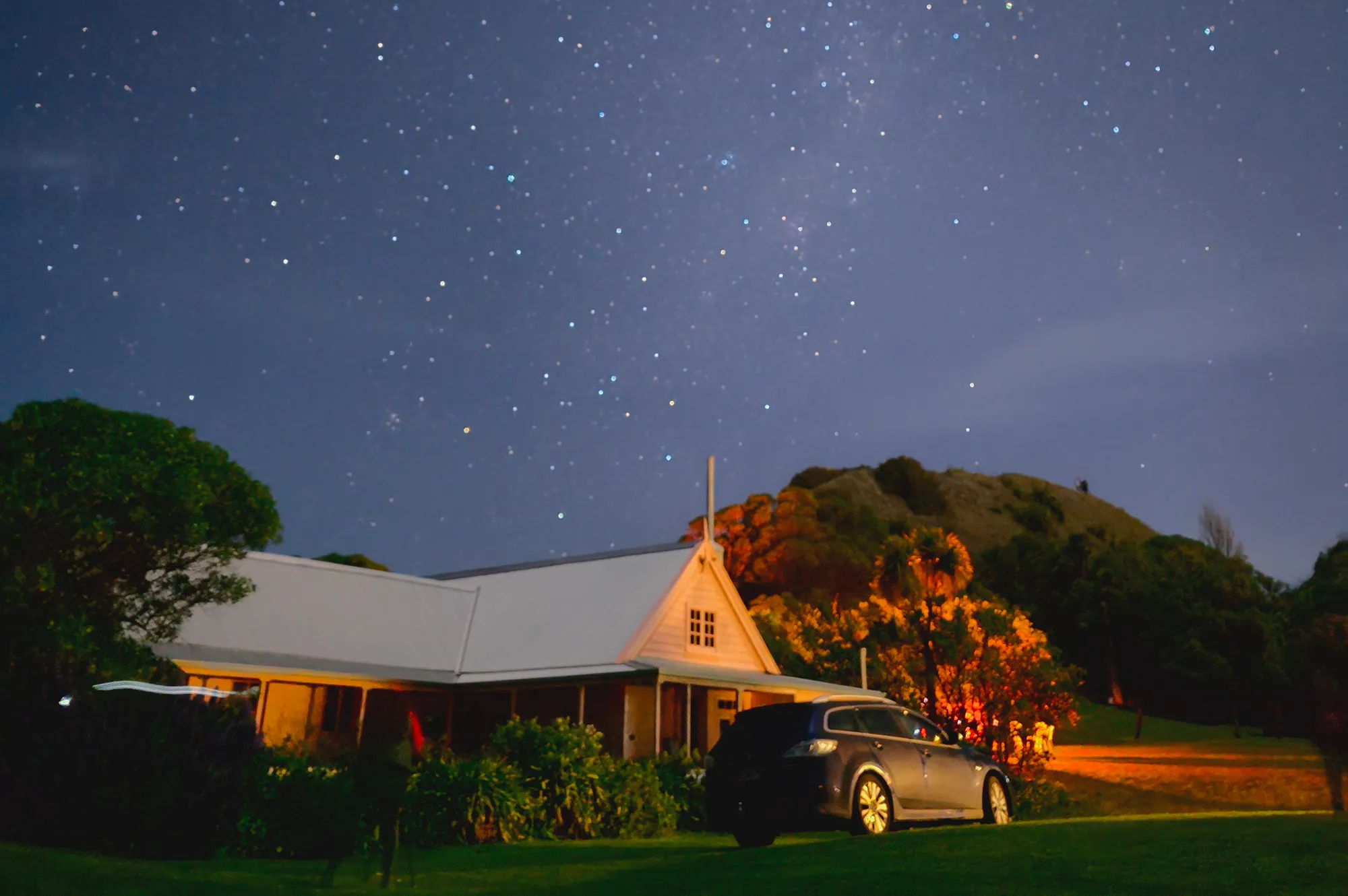 A rural farmhouse and parked car under a brilliant star-filled sky at night.