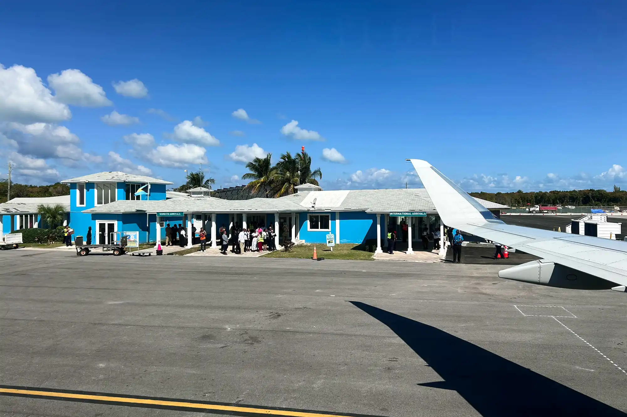 A blue airport terminal building with a crowd of travelers and an airplane wing in the foreground.