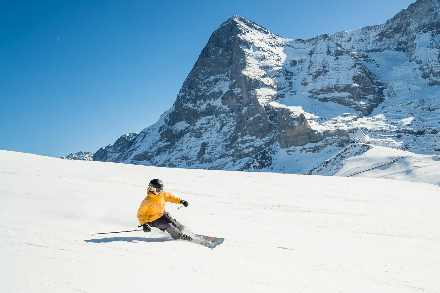 A person skiing down a wide, snowy slope with high mountains in the background.
