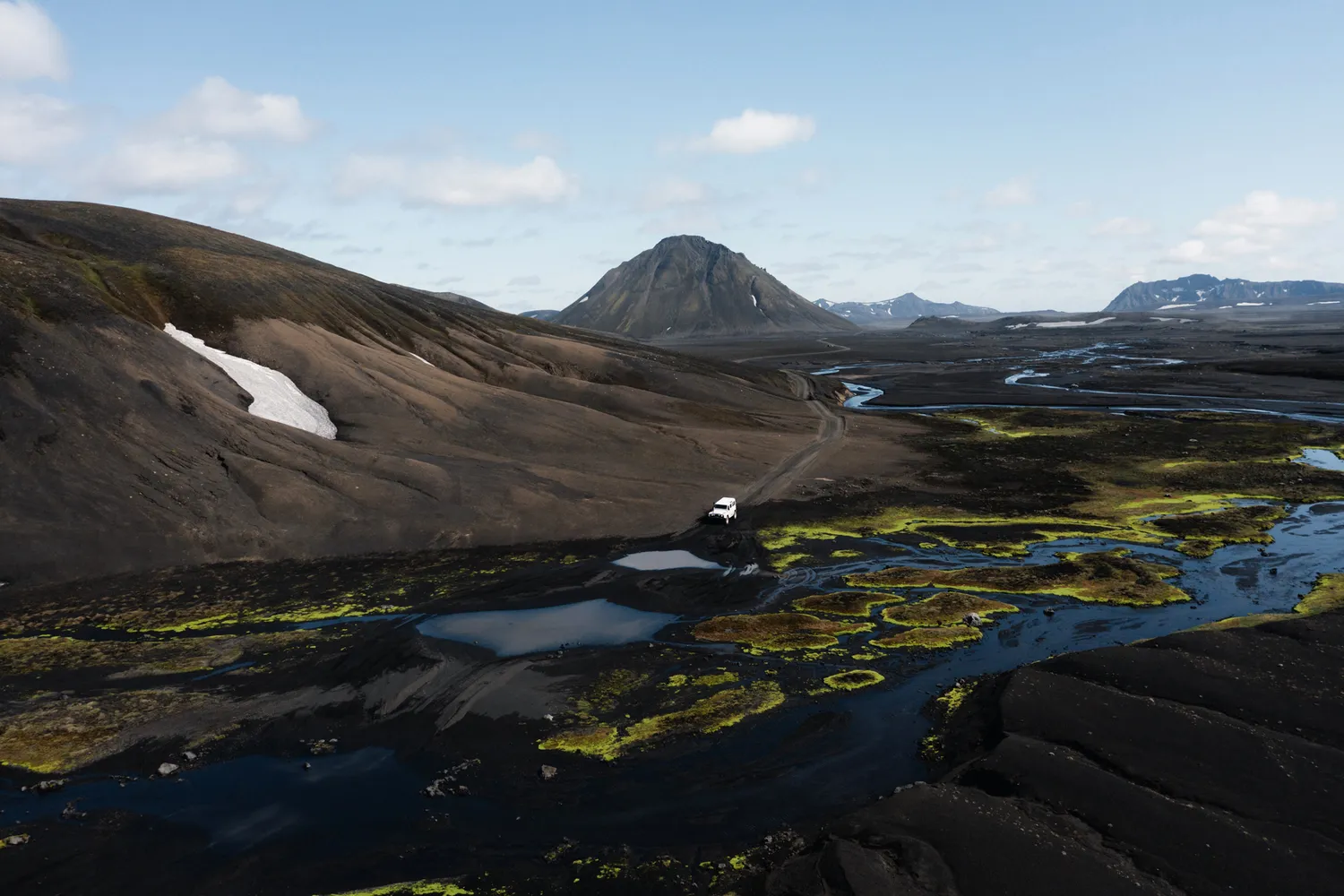 A lone car driving on a paved road through vast, green moss-covered lava fields under a cloudy sky.