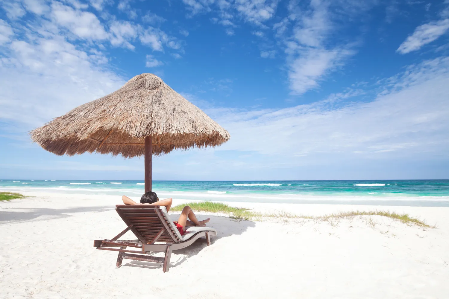 A person relaxing on a lounge chair under a traditional palapa umbrella on a white sand beach.