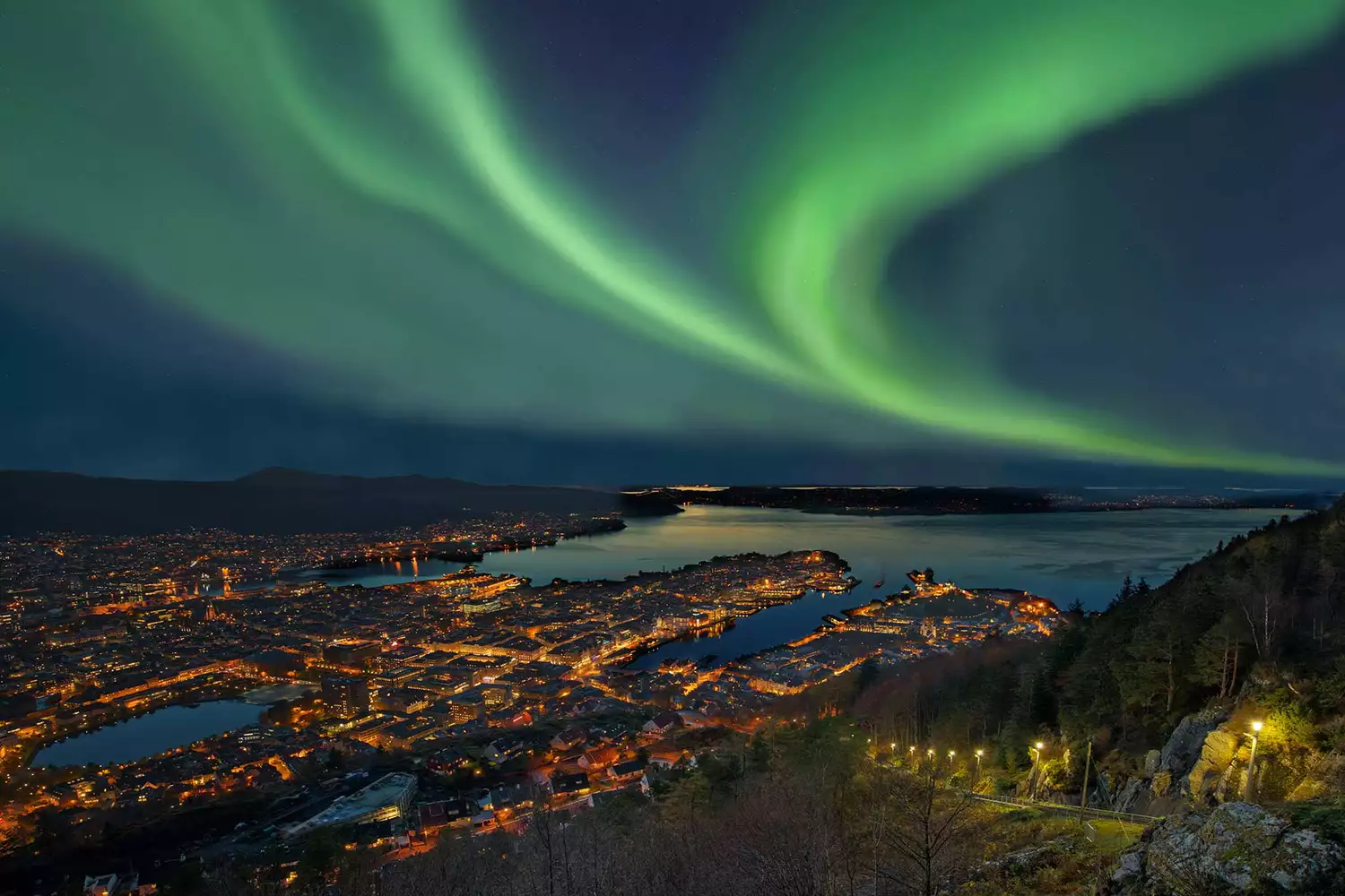 The vibrant green Northern Lights dancing over the mountains and water of Bergen, Norway.