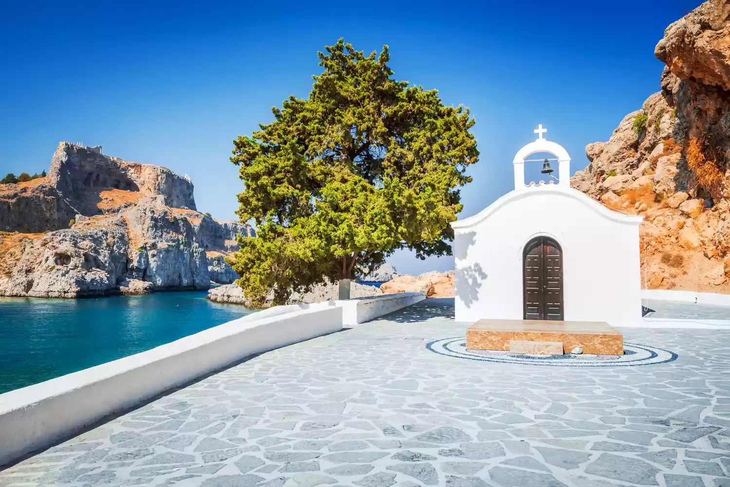A panoramic view of the white buildings of Lindos town beneath the ancient Acropolis ruins and the blue sea.