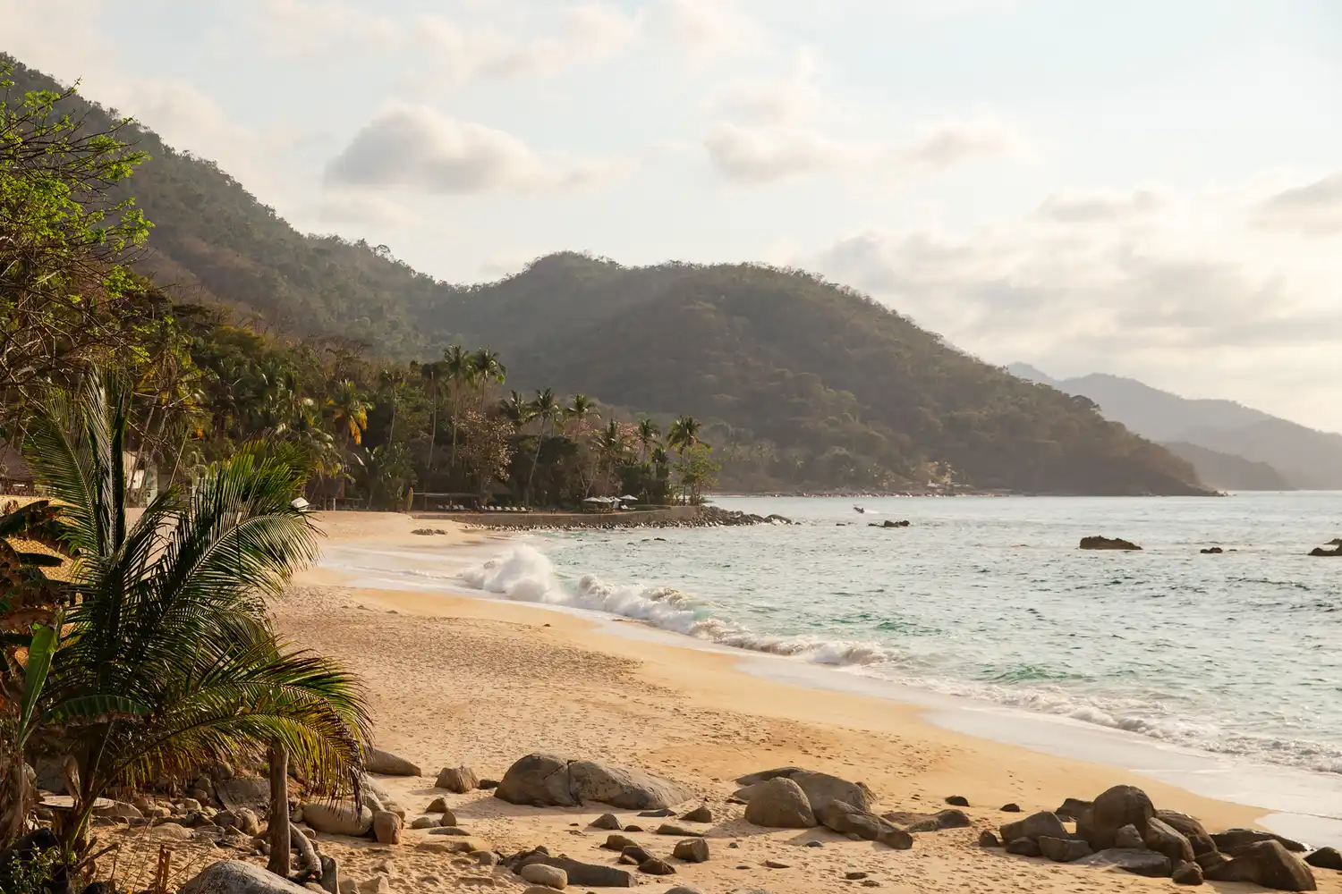 A calm, sandy beach in Puerto Vallarta with palm trees and hazy mountains in the background.