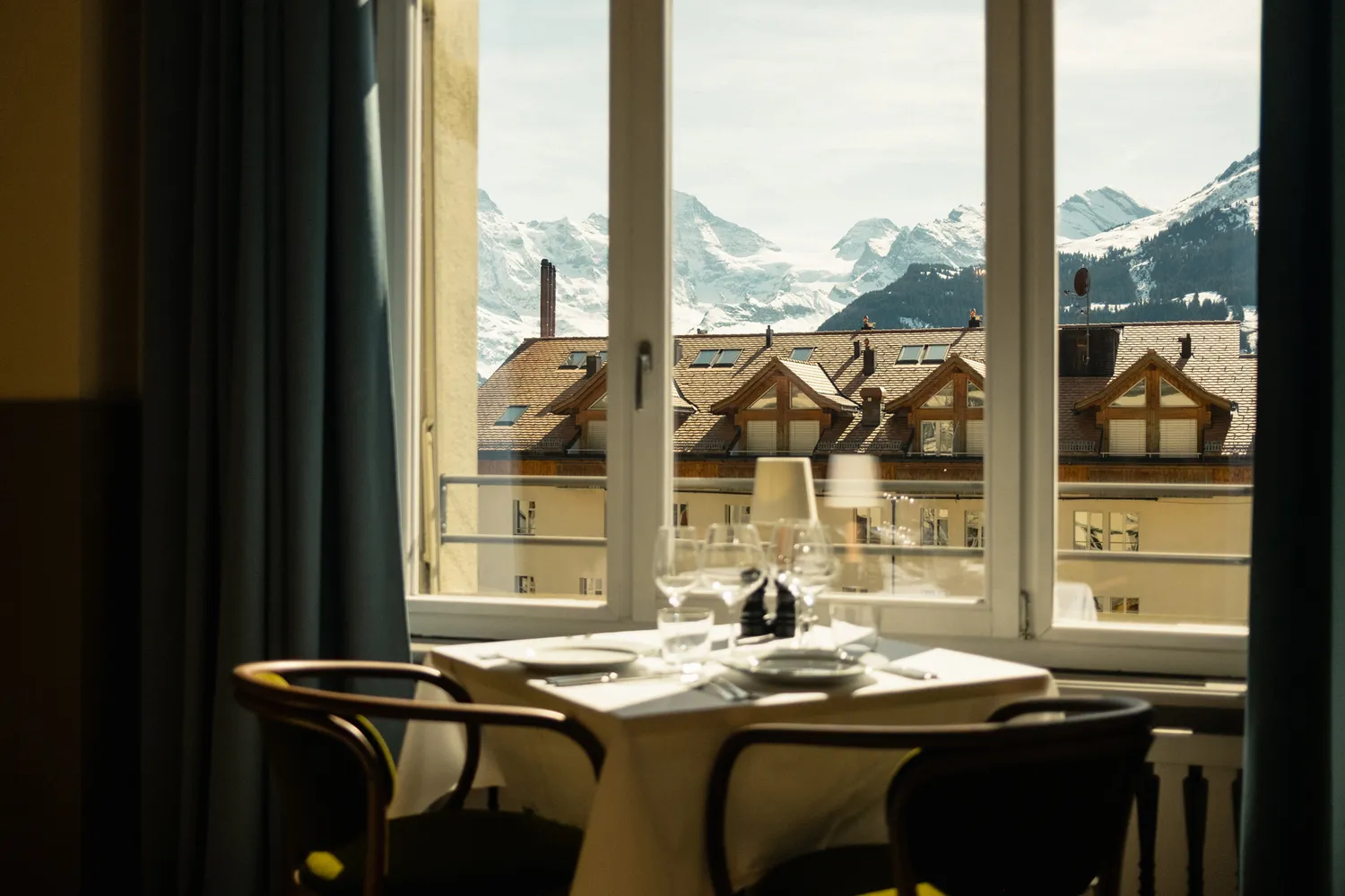 A dining table set with glassware by a window overlooking alpine rooftops and mountains.