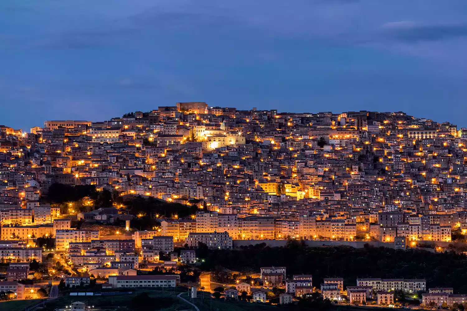A wide twilight panorama of the hill town of Gangi in Sicily, with lights glowing as the sun sets.