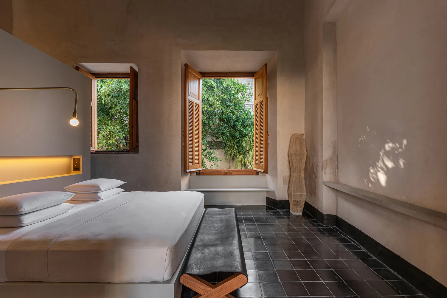 Minimalist hotel room with concrete walls, wooden furniture, and views of lush greenery.