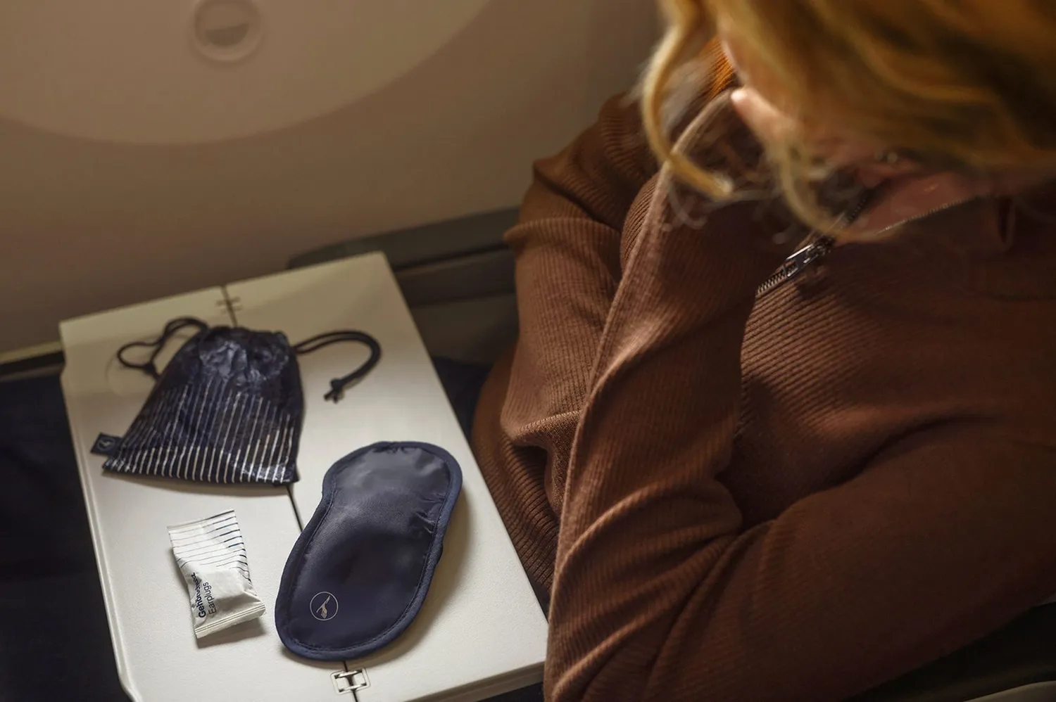 An airline passenger interacting with a new travel amenity kit containing an eye mask and earplugs on a seat tray.