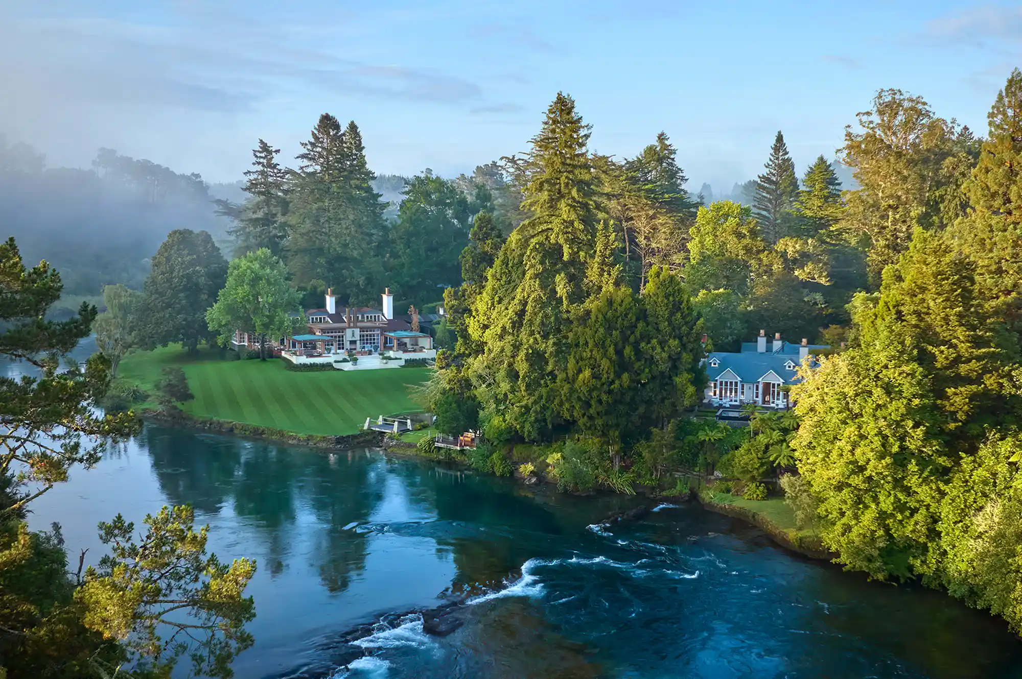 A riverside lodge with manicured green lawns surrounded by dense forest.